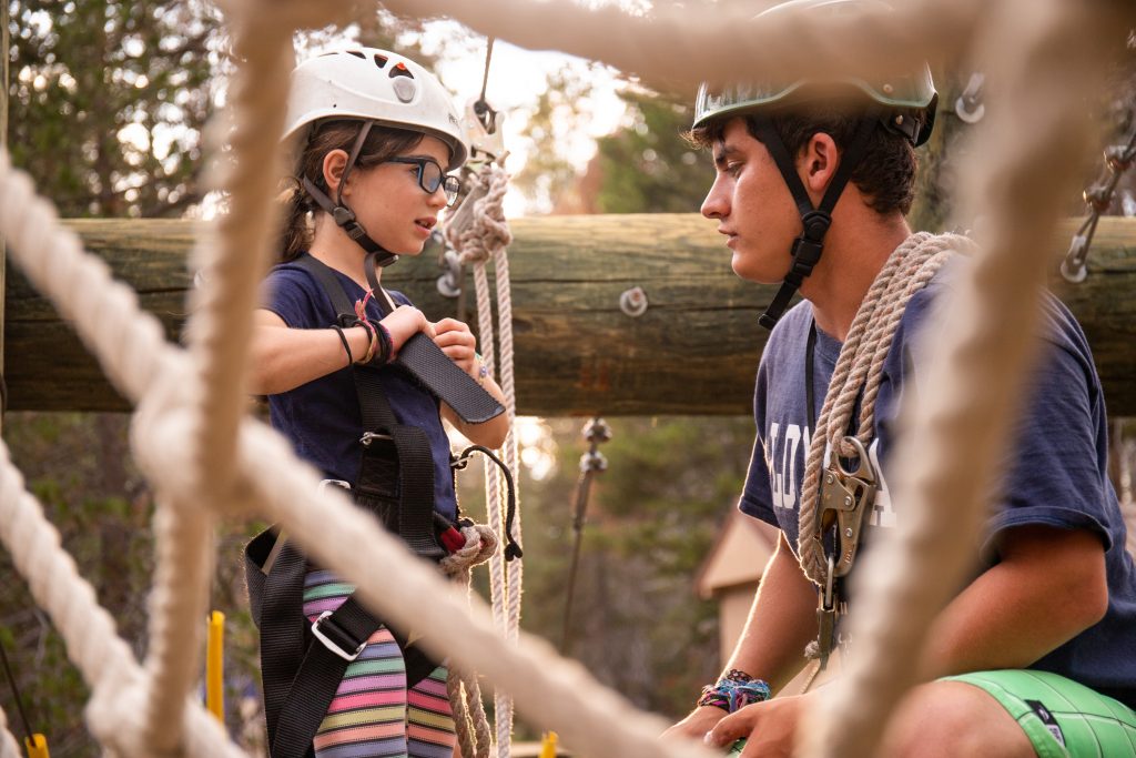 Summer Camp staff member at Gold Arrow Camp prepares a young girl to climb a cargo net